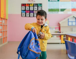 Young boy packing a blue backpack in a colorful classroom